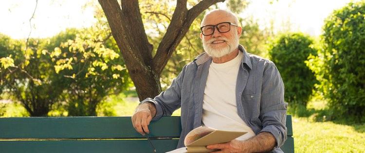 Older man sitting on an outdoor bench. He is smiling with a book open in his hand