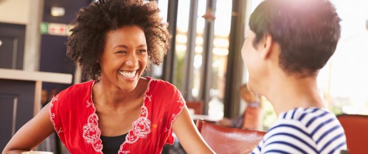 A photo of two women sit drinking coffee, smiling, and talking